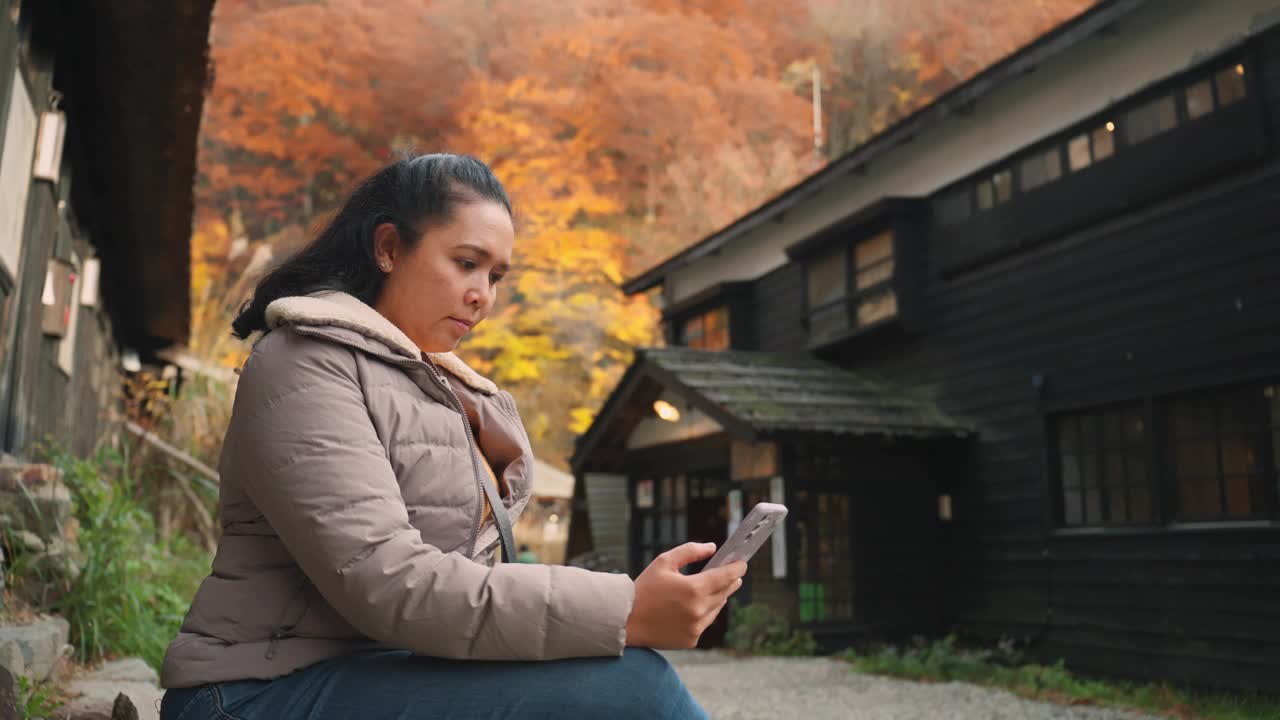 在宁静的乡村环境中，一名中年亚洲妇女坐在传统日本日式旅馆附近，背景是充满活力的秋叶，营造出一种宁静而田园诗般的氛围。视频下载