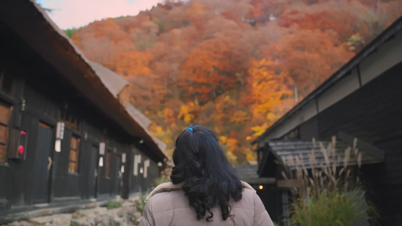 一个穿着夹克的女人的后视图，走在一条宁静的小路上，背景是一座传统的日本房子和充满活力的秋叶。这一场景凸显了日本文化的魅力和历史。视频下载