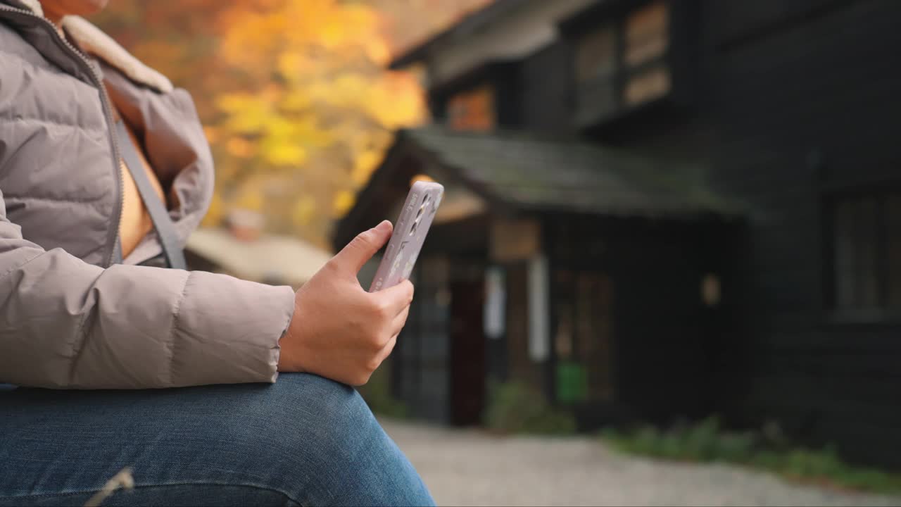 这是一个女人使用智能手机的特写镜头，她坐在户外一家传统的日本日式旅馆附近，享受着放松的时刻，也许是在欣赏周围的自然美景，强调她与宁静环境的联系。视频下载