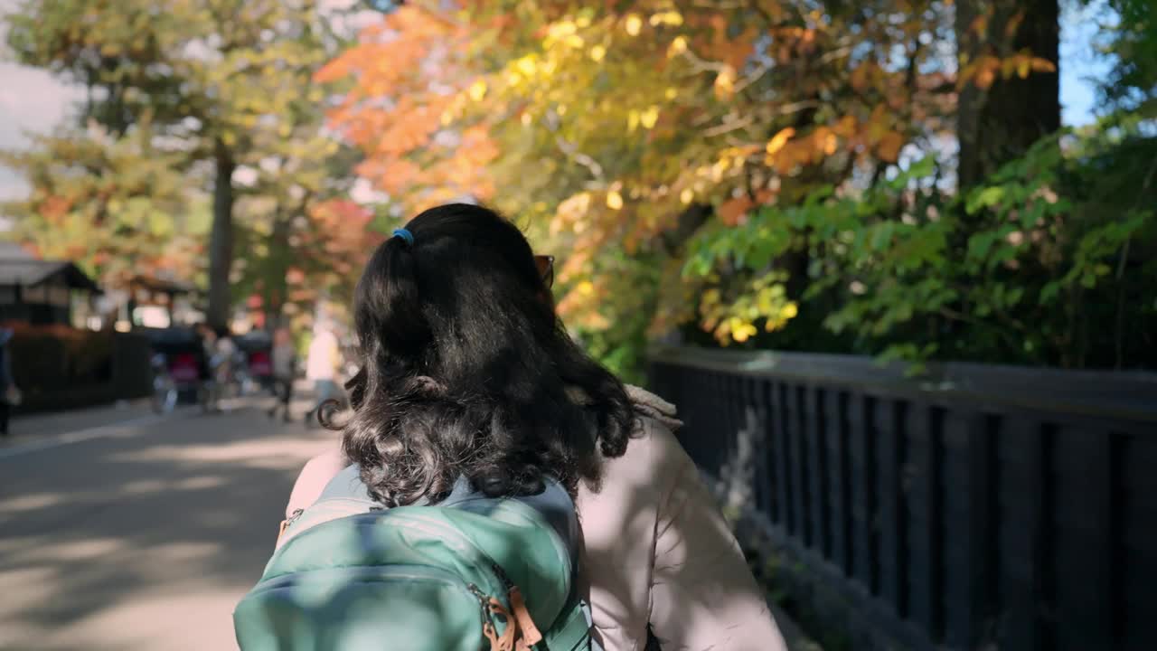 一位中年妇女的后视图，她走在一条宁静的小路上，两旁是枝繁叶茂的树木。她背着一个绿色的大背包，表明她正在徒步旅行或探索户外空间，可能是自然保护区或公园。视频下载