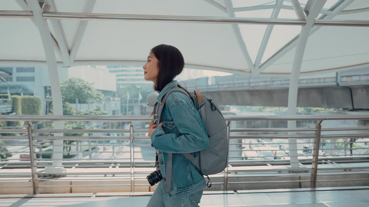 年轻的亚洲女子穿着牛仔夹克，脖子上戴着耳机，背着背包走在城市里。青春期的女性开心，笑脸盈盈。放松和享受城市生活的年轻女士视频素材