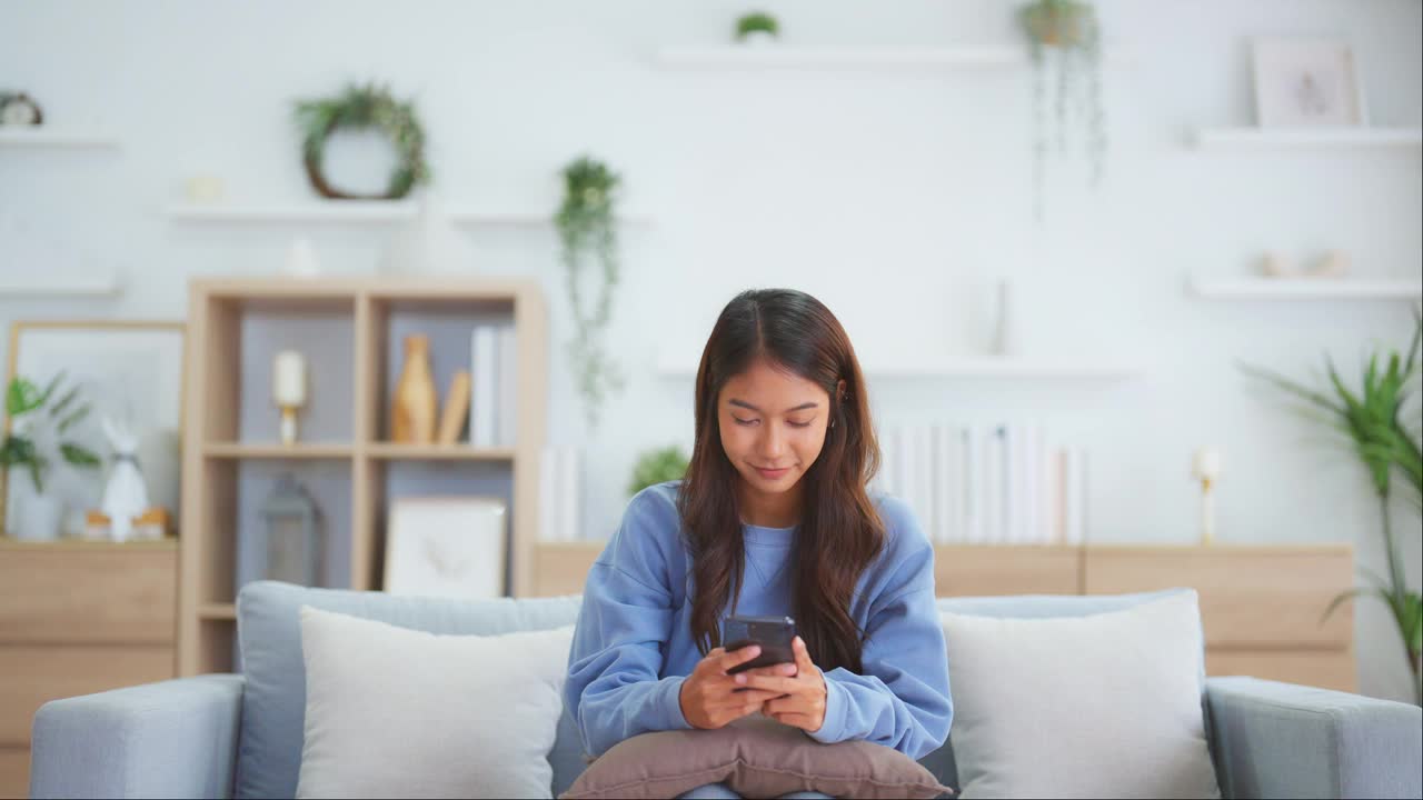 快乐的年轻亚洲女人放松在家里舒适的沙发上发短信在智能手机上，微笑的女孩使用手机，聊天在线消息，从家里网上购物视频素材