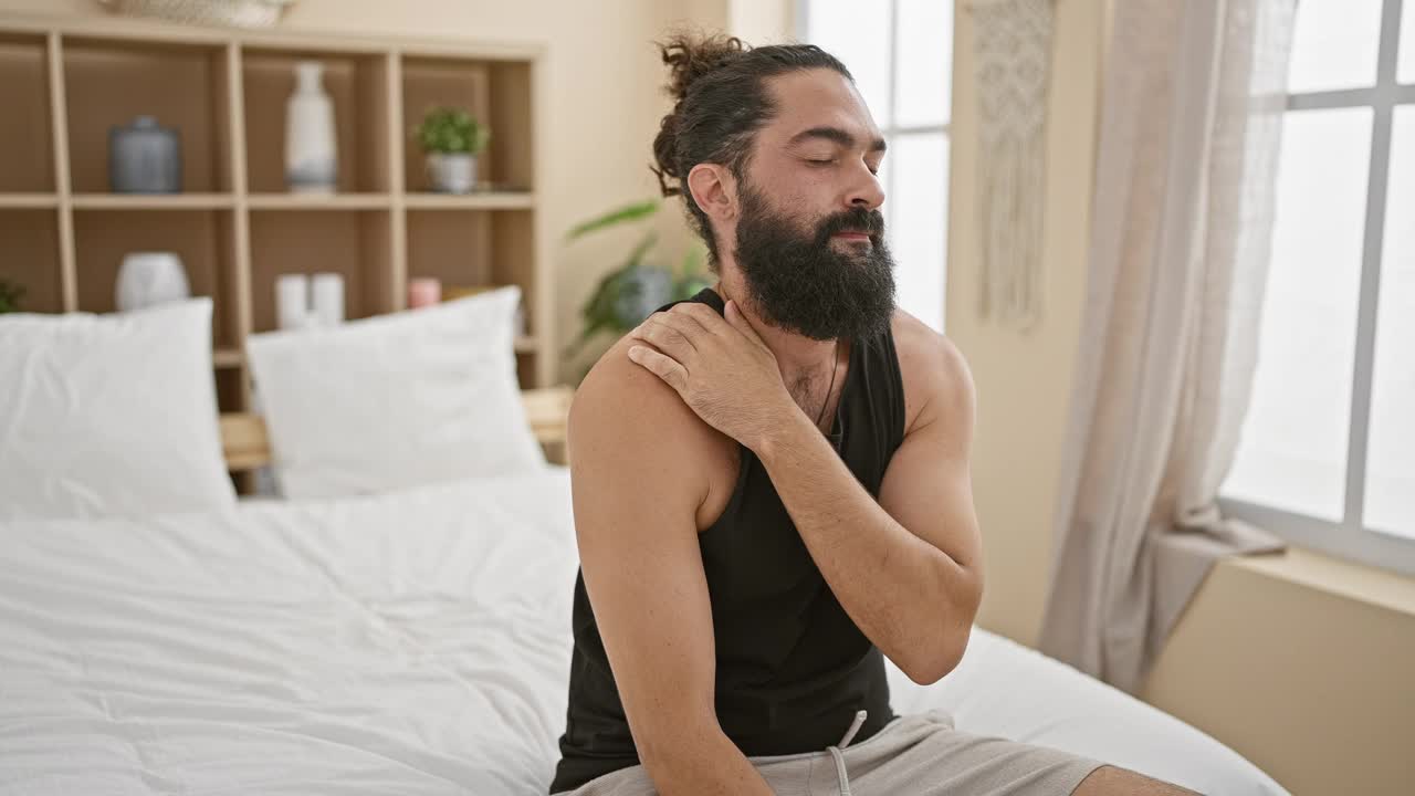 在一间光线充足的卧室里，一名大胡子男子抓着他的肩膀，露出痛苦的表情。视频素材