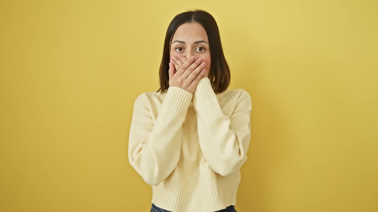 震惊和震惊，年轻漂亮的西班牙裔女子穿着毛衣，孤立在黄色背景上，在一个令人惊讶的错误之后，用手捂住嘴，无言的恐惧。视频素材