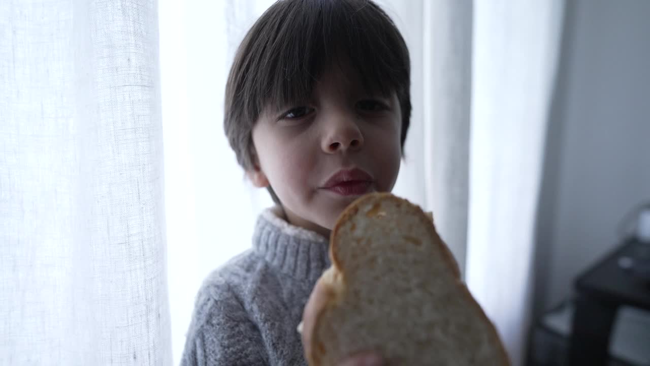 小男孩在窗边咬了一口三明治。背光下5岁的白人小孩拿着面包吃零食的场景视频素材