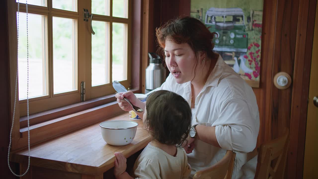 一位妇女正在给孩子喂早餐。孩子坐在桌子旁，面前放着一碗麦片粥。这个场景是温暖和滋养的视频素材