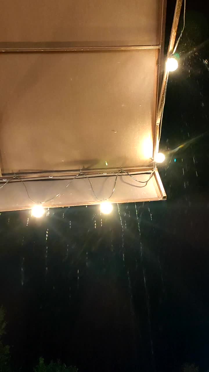 雨滴从屋顶滴下，晚上有发光的灯笼。在黑暗的夜晚，雨滴从覆盖着灯泡花环的边缘流淌。雨滴飞溅。垂直视频素材