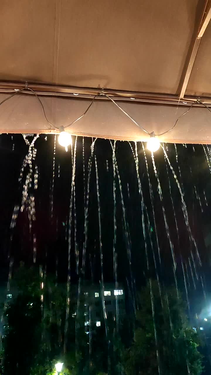 雨点从屋顶滴下，晚上灯笼发光。在黑暗的夜晚，雨水从覆盖着灯泡花环的边缘流淌。大雨飞溅。垂直视频素材