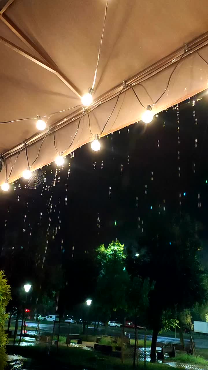 雨点从屋顶滴下，晚上灯笼发光。在黑暗的夜晚，雨水从覆盖着灯泡花环的边缘流淌。城里下大雨。垂直视频素材