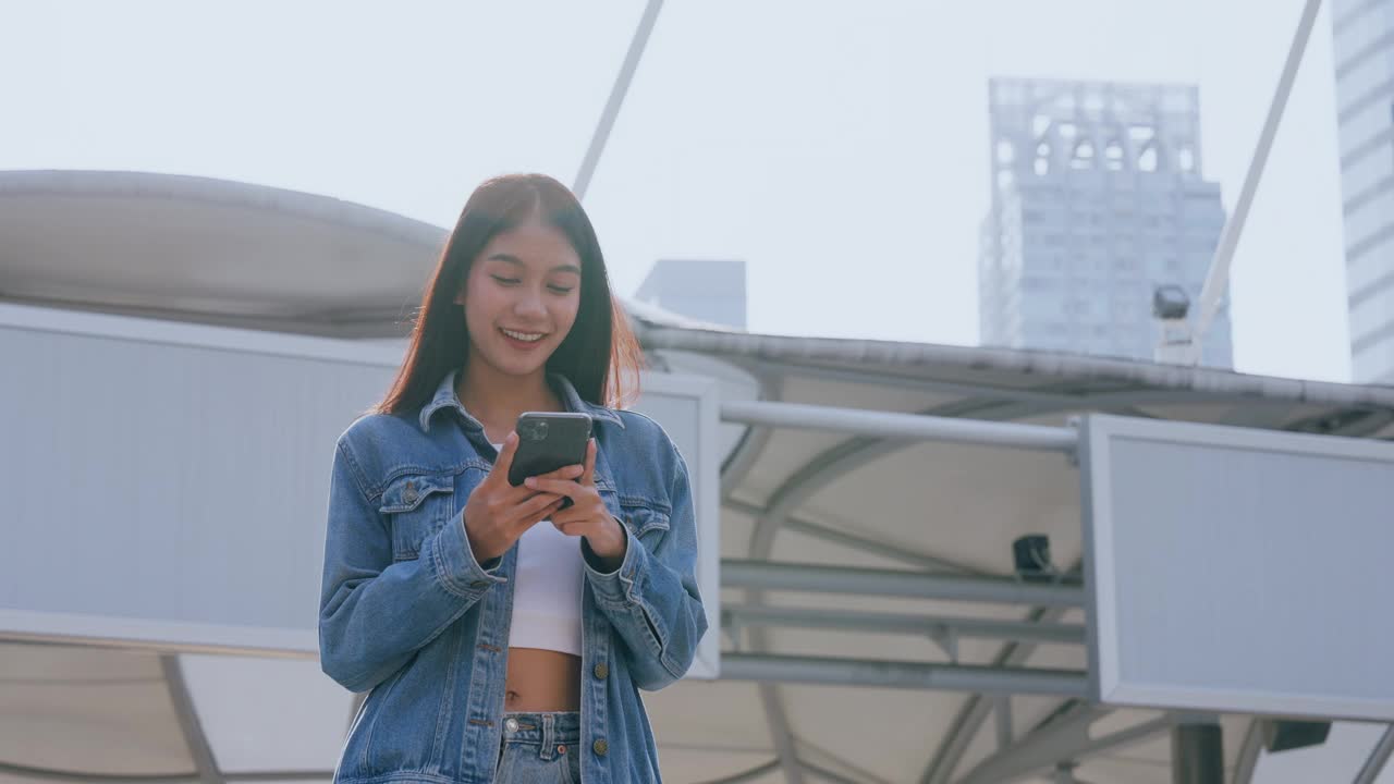 年轻的亚洲女子穿着牛仔裤，夹克，衣服，在晴朗的日子里走在城市的街道上，用智能手机发短信视频素材