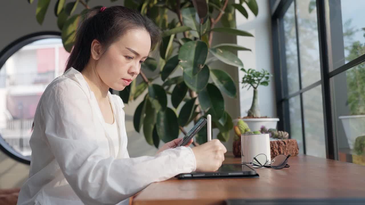 一个女人坐在桌子旁，手里拿着平板电脑和手机视频素材