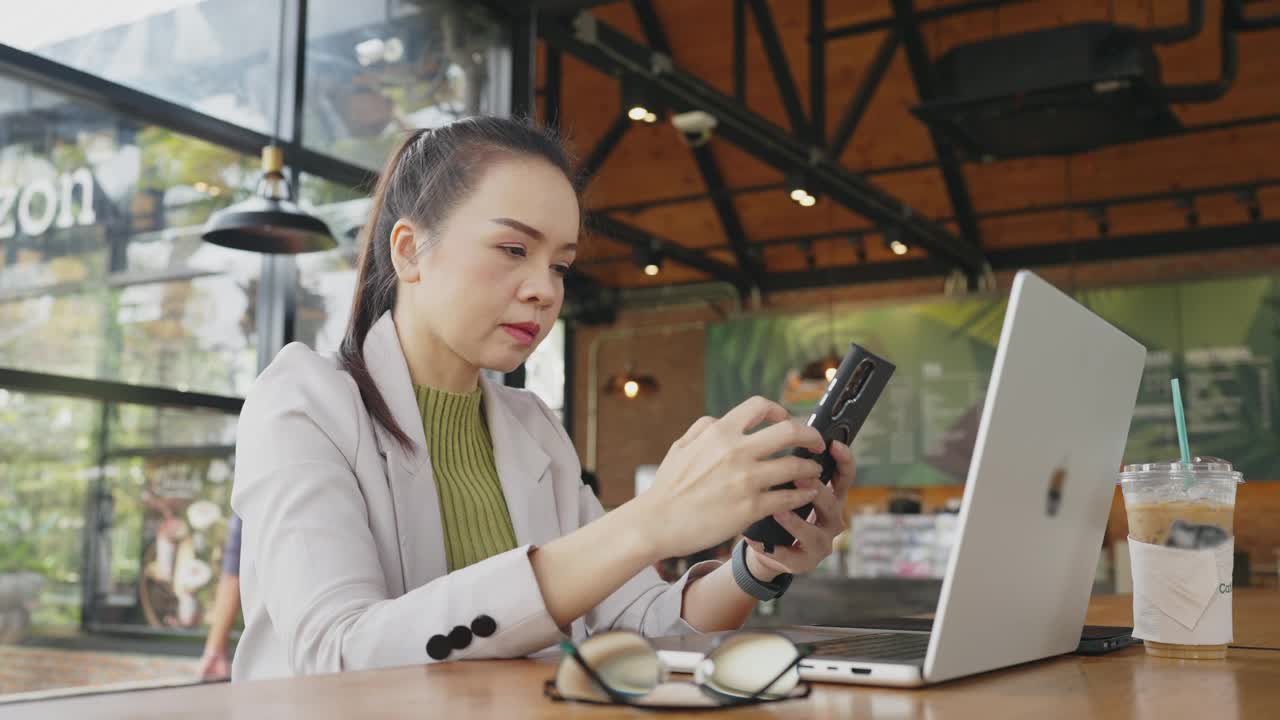 一个女人坐在桌子旁，手里拿着一台笔记本电脑和一部手机视频素材