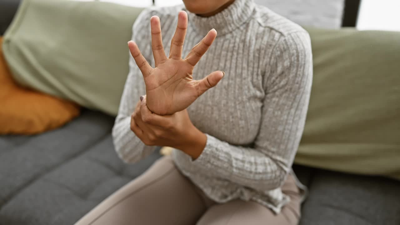 一个穿着高领毛衣、指甲修剪整齐的女人坐在室内的沙发上，表现出双手疼痛或不适。视频素材