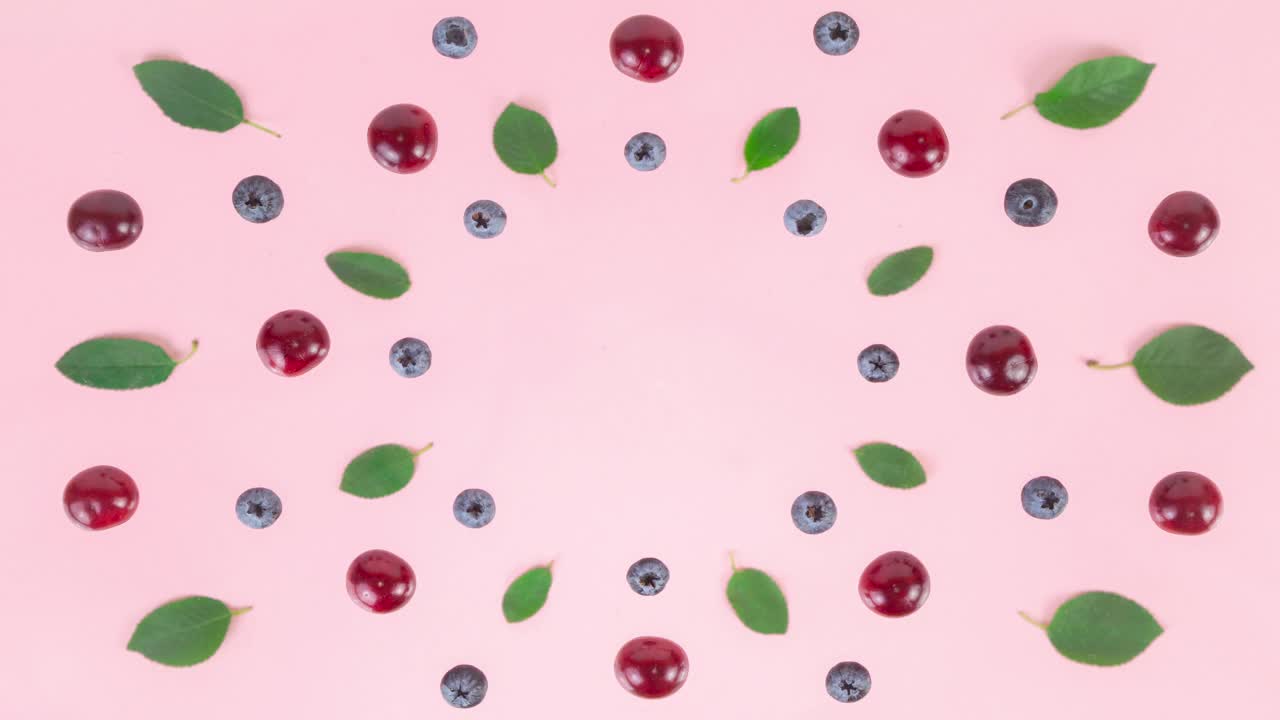 成熟的樱桃、蓝莓和绿叶在粉红色的背景上排成一个圆圈。视频下载