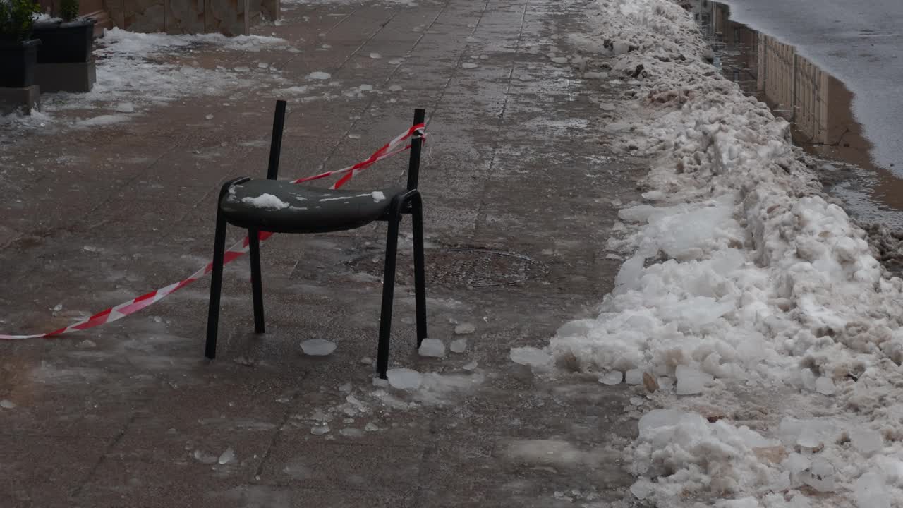 一张椅子坐在积雪的人行道上，旁边是一座建筑物，附近有水流视频素材