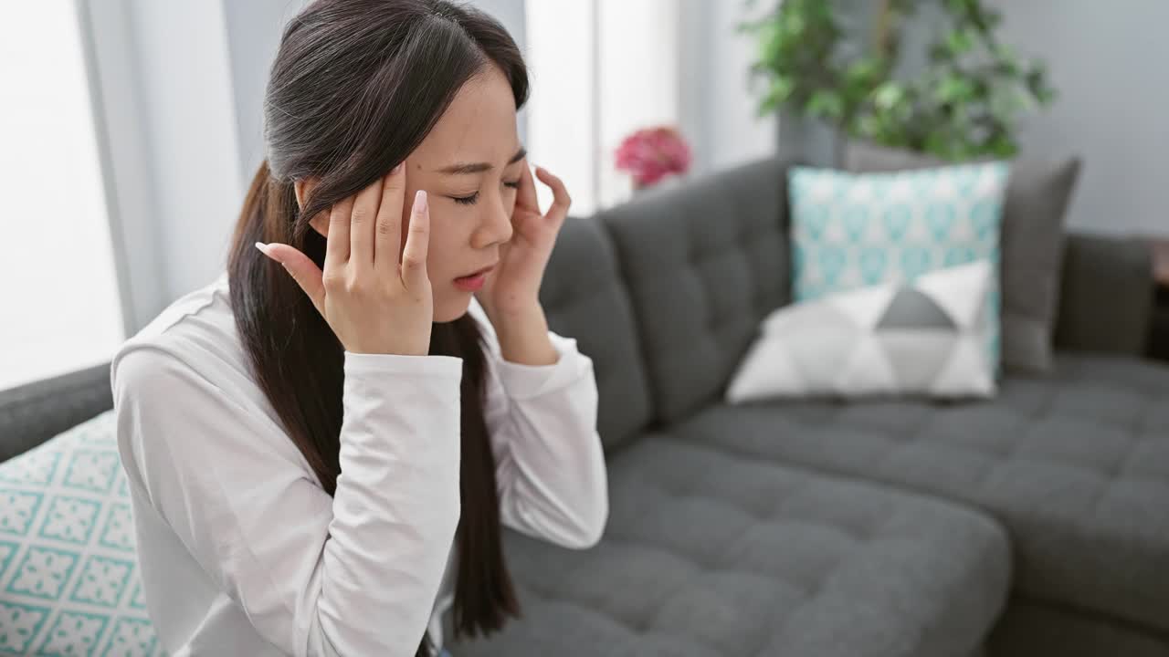 一位年轻的亚洲女性在室内遇险，在一个现代化的客厅里抱着她的头。视频素材