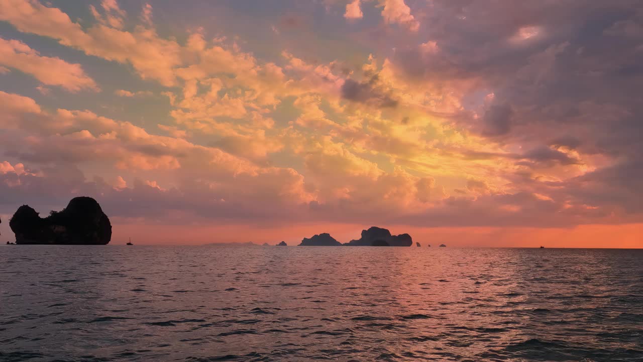 在平静的海面上，令人惊叹的日落将天空描绘成充满活力的橙色、粉色和黄色。远处岛屿和船只的剪影为宁静的景色增添了深度。泰国视频素材