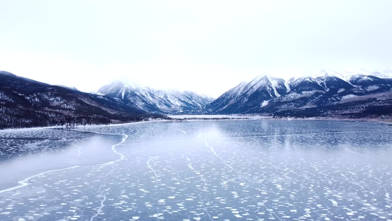 在冬天远离白雪皑皑的山脉，在冰冻的湖面上高空飞行视频素材