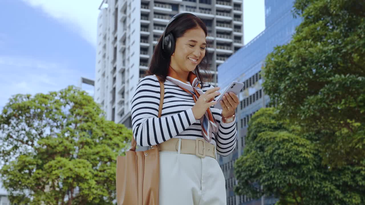 女人带着耳机和手机在城市里微笑，在街上唱韩国流行歌曲或听网络广播。亚洲人，智能手机和快乐的音频流订阅，用于旅行，跳舞和通勤视频素材