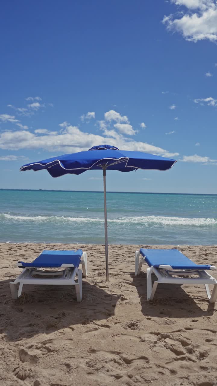 一个宁静的海滩场景，蓝色的遮阳伞和配套的日光浴床在晴朗的天空下俯瞰海洋视频素材