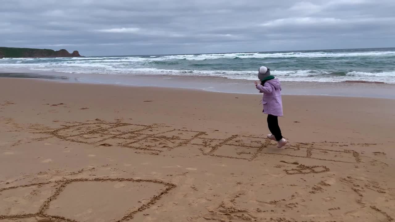 蹒跚学步的小女孩跳跳房子，海景沙滩上的经典。与家人和孩子的日常户外活动。玩户外游戏。视频下载