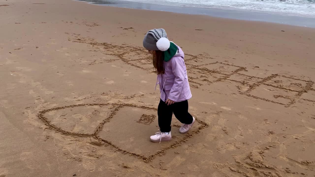 蹒跚学步的女孩在沙滩上画画，可以看到海景。与家人和孩子的日常户外活动。玩户外游戏。视频下载