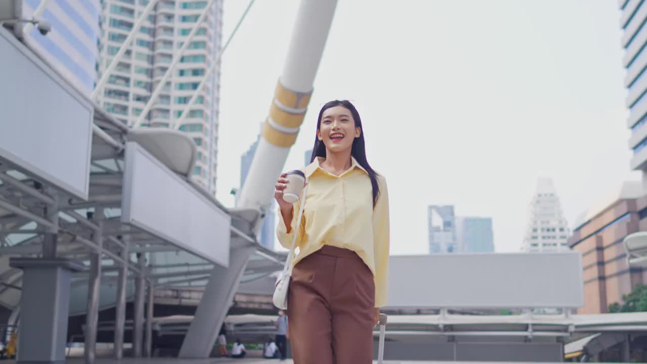 亚洲年轻美丽的女商人在现代城市户外散步。漂亮的女员工拿着一杯热咖啡，拎着行李，在市中心散步，感到快乐和自信。视频素材