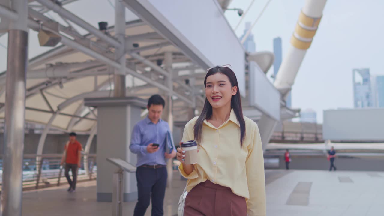 亚洲年轻美丽的女商人在现代城市户外散步。迷人的年轻员工女孩感到快乐和自信，同时拿着一杯热咖啡在镇中心散步。视频素材