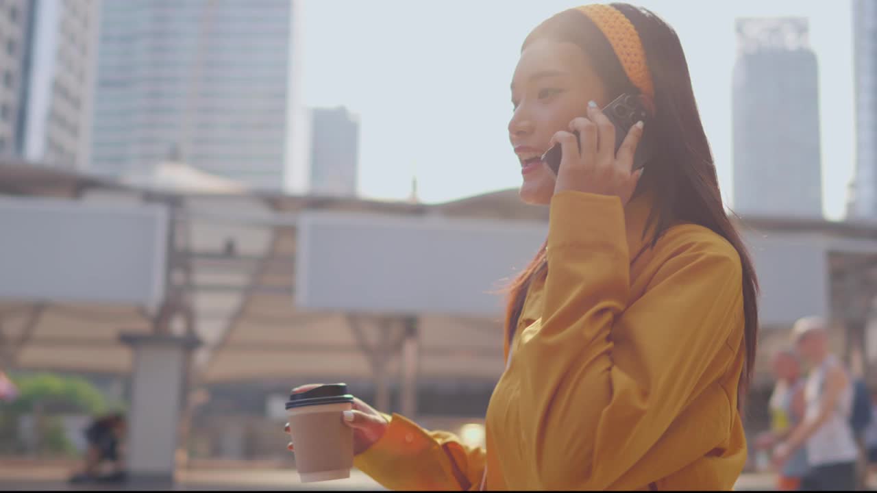 亚洲美女在城市户外使用数字智能手机交流。迷人的女商人端着一杯热咖啡，一边打电话一边走在镇中心。视频素材