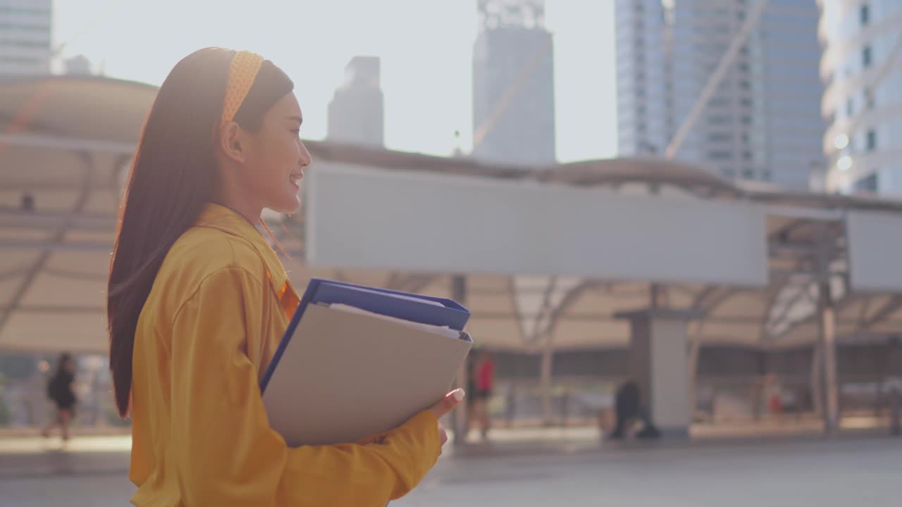 亚洲年轻美丽的女商人在现代城市户外散步。迷人的女员工感到快乐和自信，拿着剪贴板，走在镇中心的办公室外面视频素材