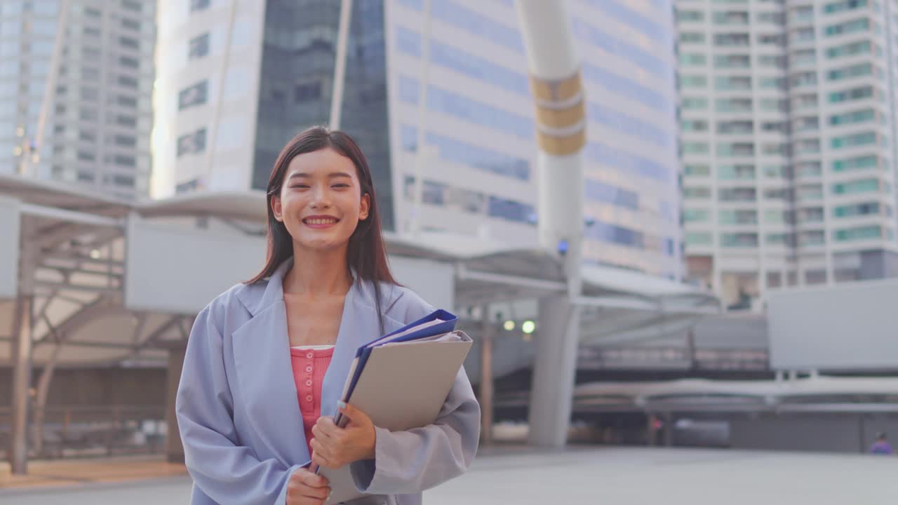 站在现代都市户外的亚洲年轻女商人肖像。迷人的女员工感到快乐和自信，拿着剪贴板，看着镜头在镇中心。视频素材