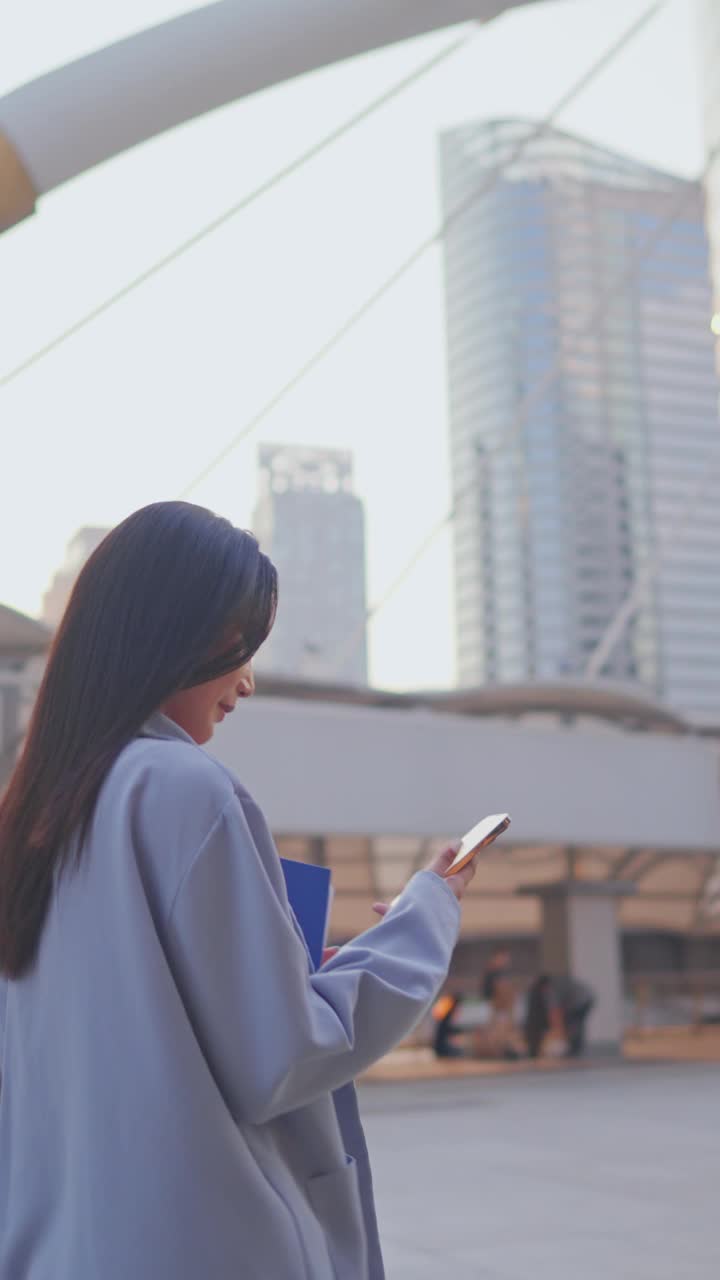 亚洲美女在城市户外使用数字智能手机交流。迷人的女商人拿着工作剪贴板，边走边打电话，走在镇中心视频素材