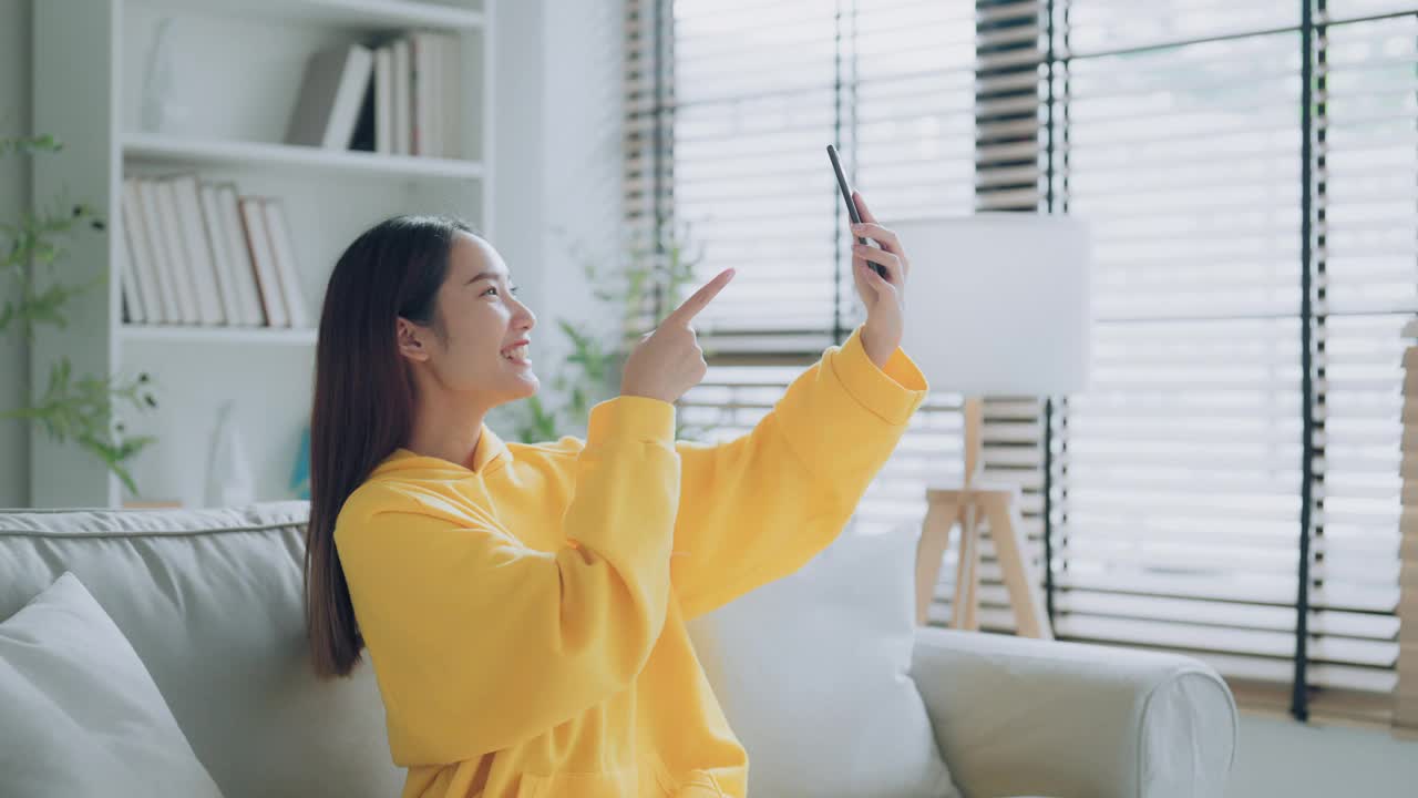 快乐的年轻亚洲女性在家里放松。坐在沙发上拿着手机的女性微笑着。女孩用视频打电话给朋友视频素材