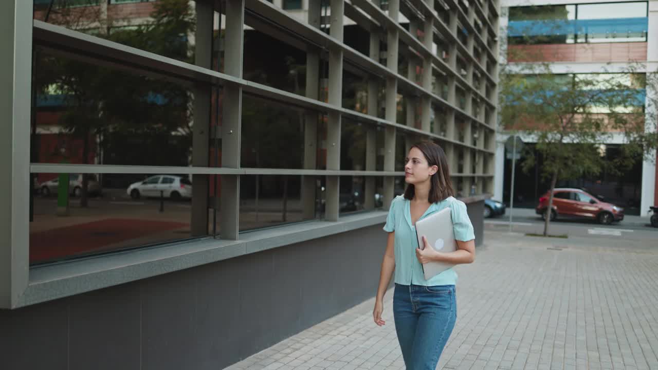 穿着休闲装的商务女性走在城市里，手里拿着笔记本电脑走进办公室开始一天的工作视频素材