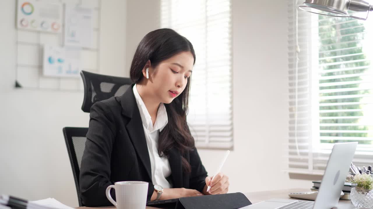 这位勤奋的年轻亚洲女商人在她的平板电脑上做笔记，在她的办公桌上用笔记本电脑阅读与项目相关的文件视频素材