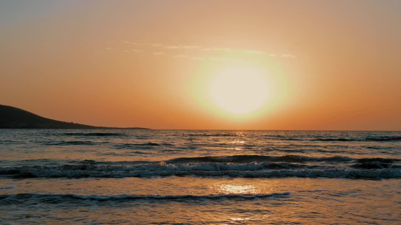 太阳正落在海面上，在水面上投下温暖的光芒。天空是橙色和粉色的混合色调，营造出一种宁静祥和的氛围。海浪轻轻地拍打着海岸视频下载