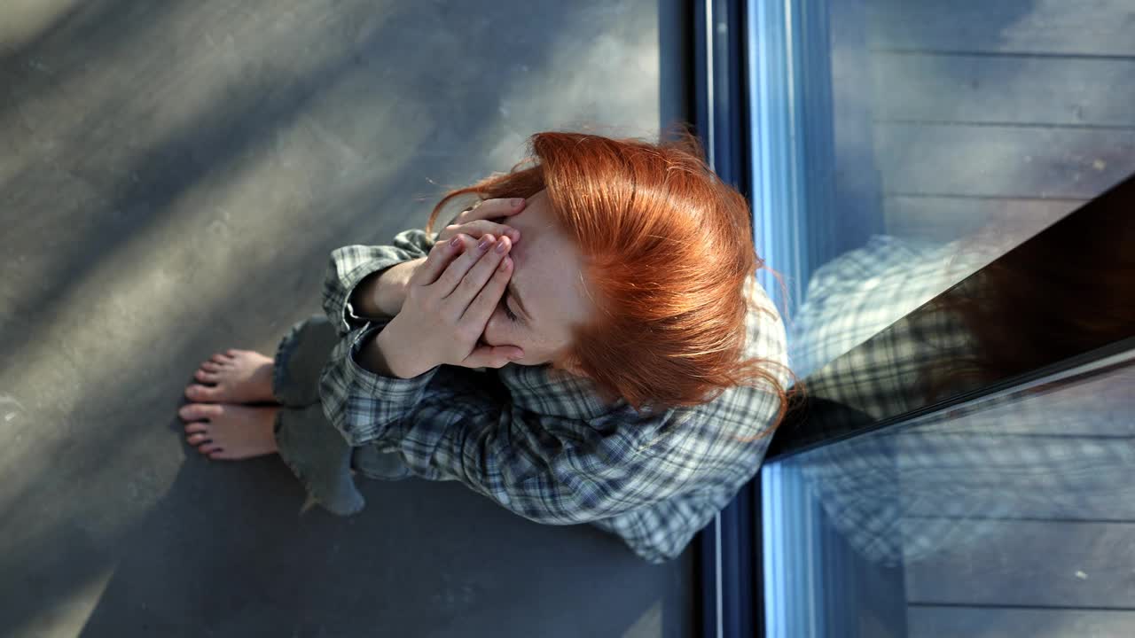 俯视图，悲伤的女人为坏消息哭泣，悲伤或抑郁的损失，心理健康或家里的问题，坐在窗边。情绪化的红发女人在流泪视频素材