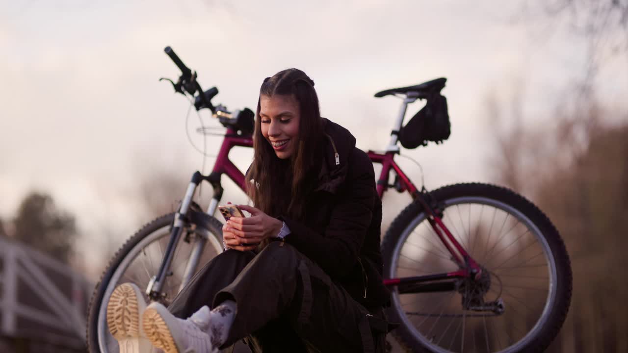 这张照片拍摄于日落时分，一位年轻女子坐在风景优美的户外自行车旁，用智能手机自拍。视频素材