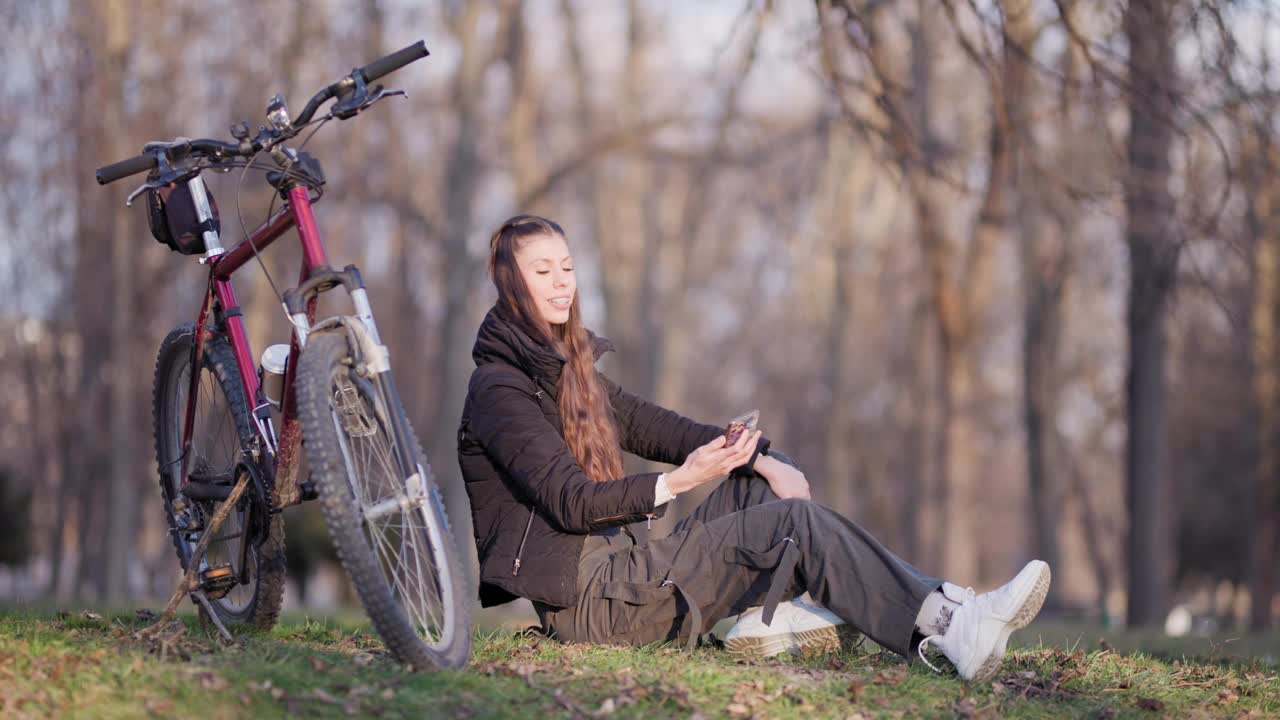 在一个阳光明媚的日子里，一名女子在公园里的自行车旁放松，用手机自拍了一张快乐的瞬间。视频素材