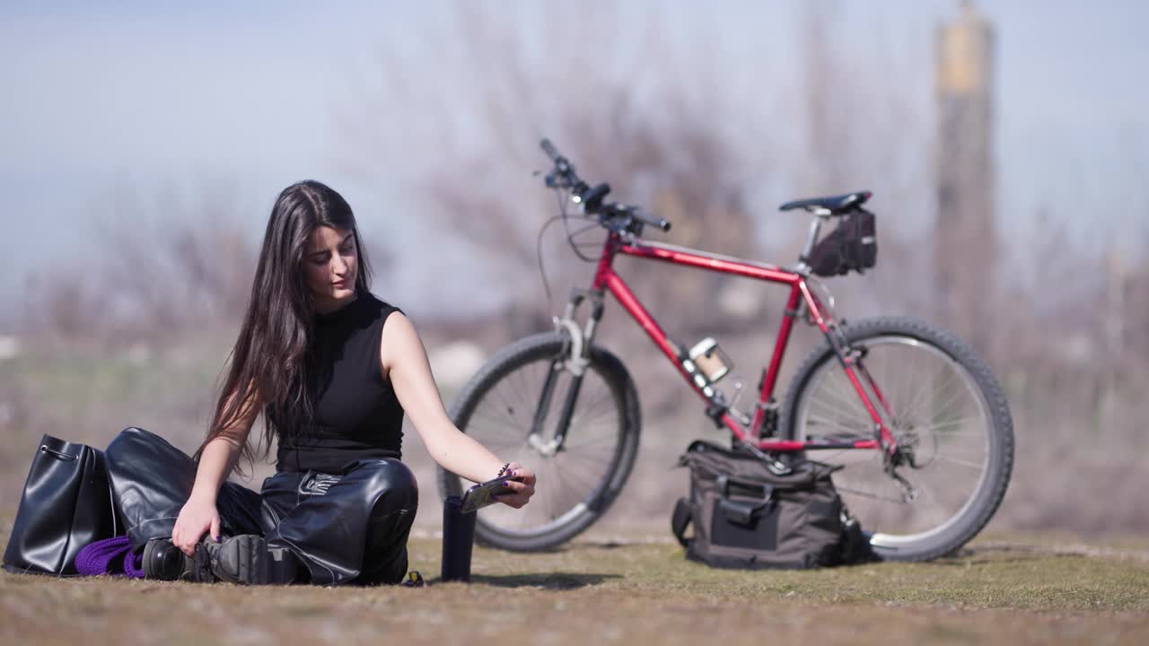 一位年轻的女自行车手坐在户外的地上，在她的红色自行车和装备包旁边自拍。视频素材