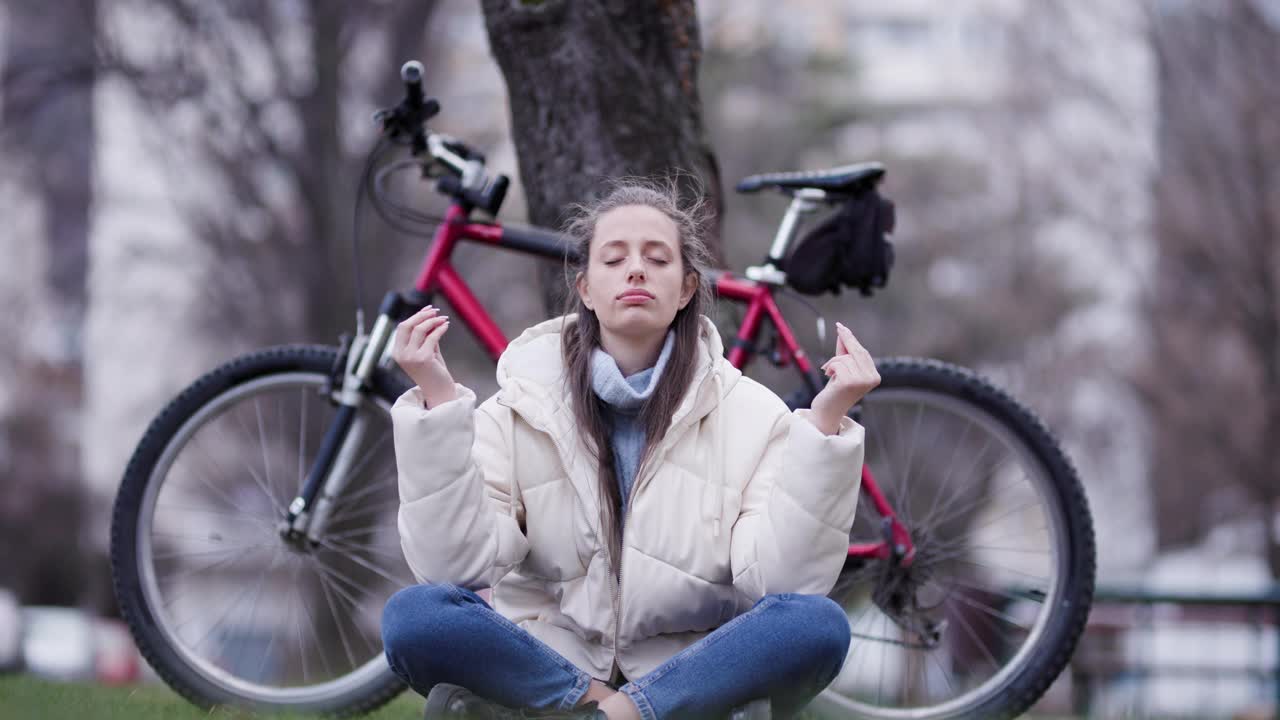 在一个宁静的公园里，一名年轻女子在练习冥想，她盘腿坐着，自行车停在旁边，在大自然中传达一种宁静和正念的感觉。视频素材