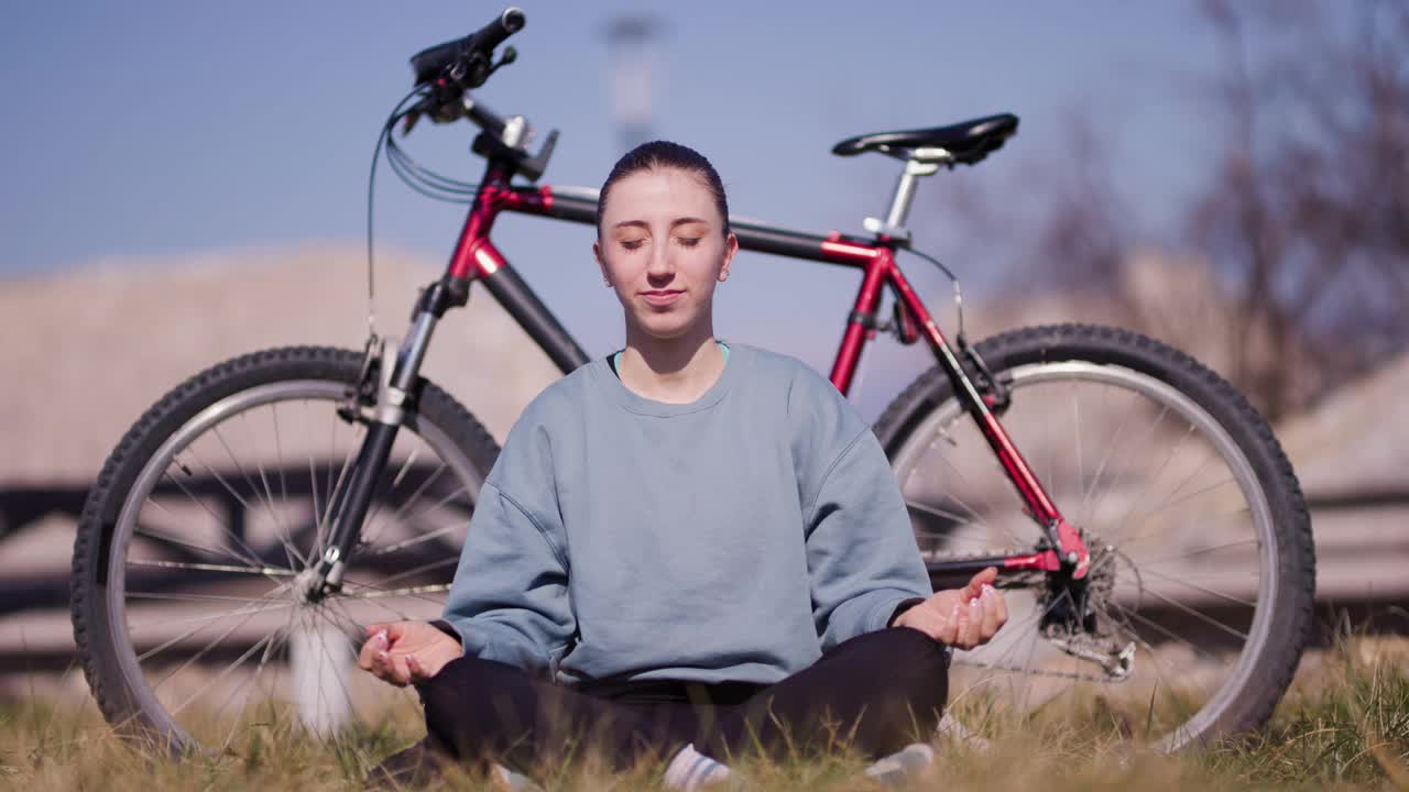 一个穿着休闲装的快乐的年轻女子坐在草地上，旁边是她的红色自行车，在晴朗的蓝天下微笑着伸展身体。视频素材