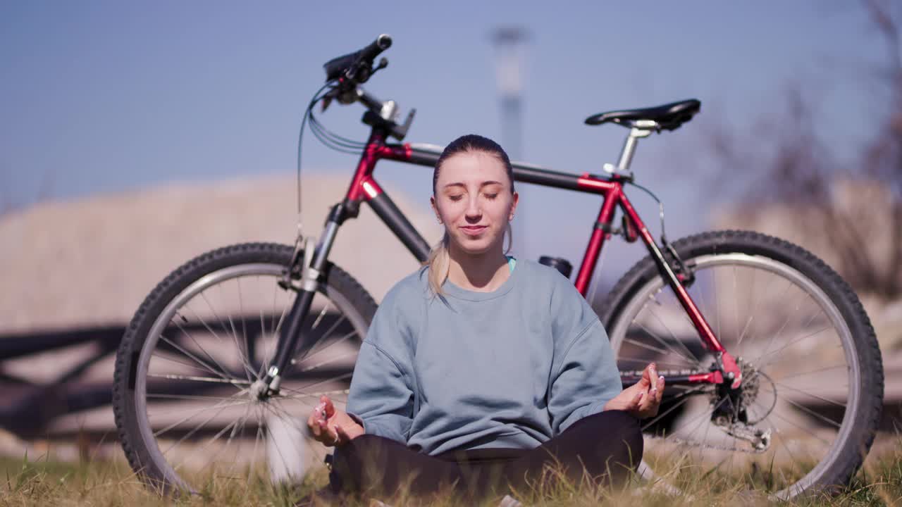 一个穿着休闲装的快乐的年轻女子坐在草地上，旁边是她的红色自行车，在晴朗的蓝天下微笑着伸展身体。视频素材