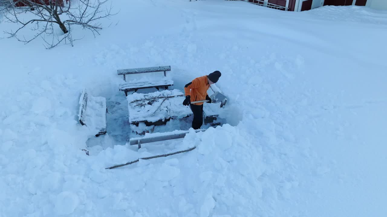 一名身穿橙色夹克的男子站在被积雪掩埋的长凳旁清理积雪。侧视图航拍视频，雪下烧烤的地方视频下载