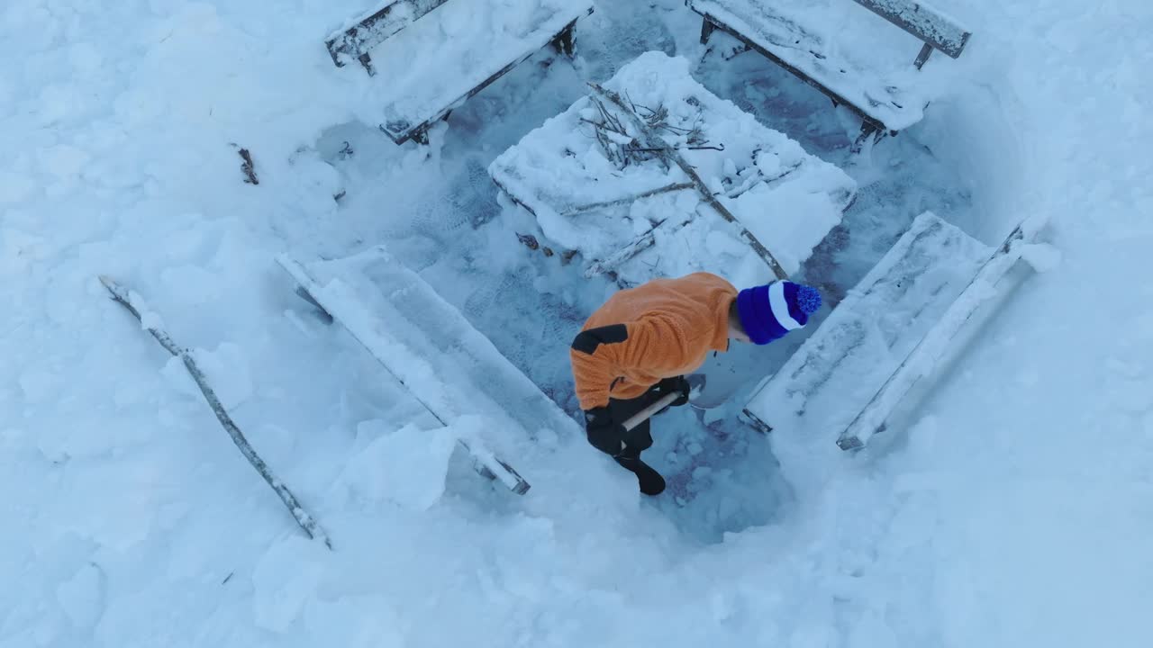 一名身穿橙色夹克的男子站在被积雪掩埋的长凳旁清理积雪。侧视图航拍视频，雪下烧烤的地方视频下载