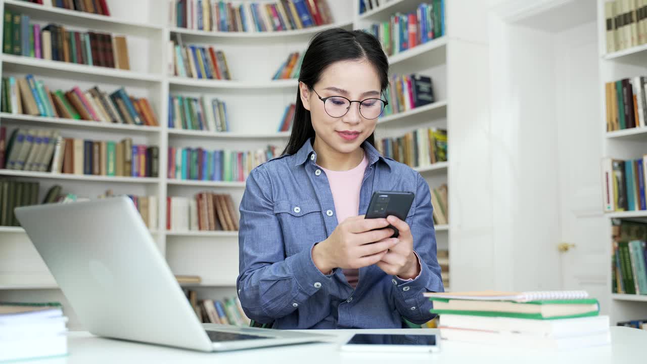 高兴兴奋的亚洲女学生庆祝成功阅读伟大的新闻，而坐在校园图书馆空间的电话。视频素材