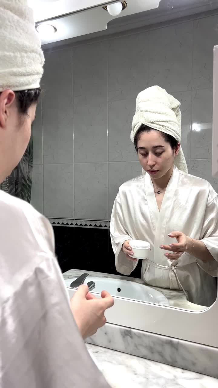 垂直视频的肖像，年轻美丽的女人在浴巾微笑，在她的脸上涂抹身体霜。视频下载