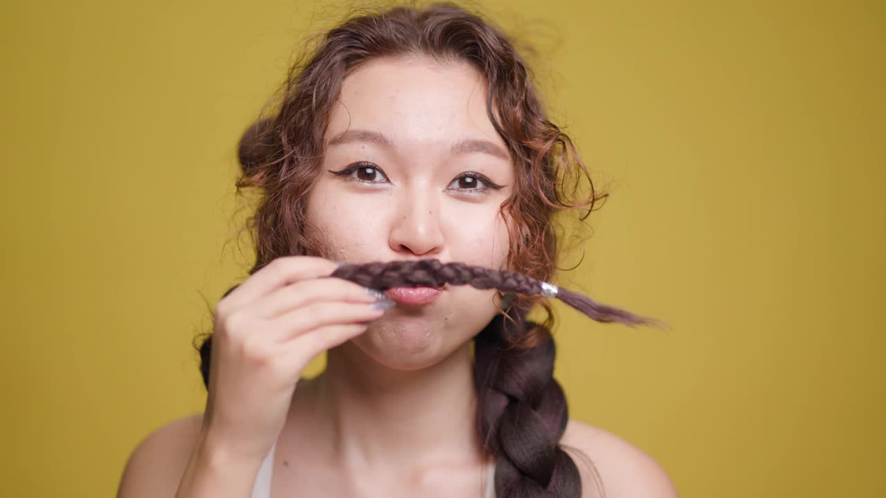 俏皮的年轻女子用她的辫子在黄色的背景下做着小胡子视频素材