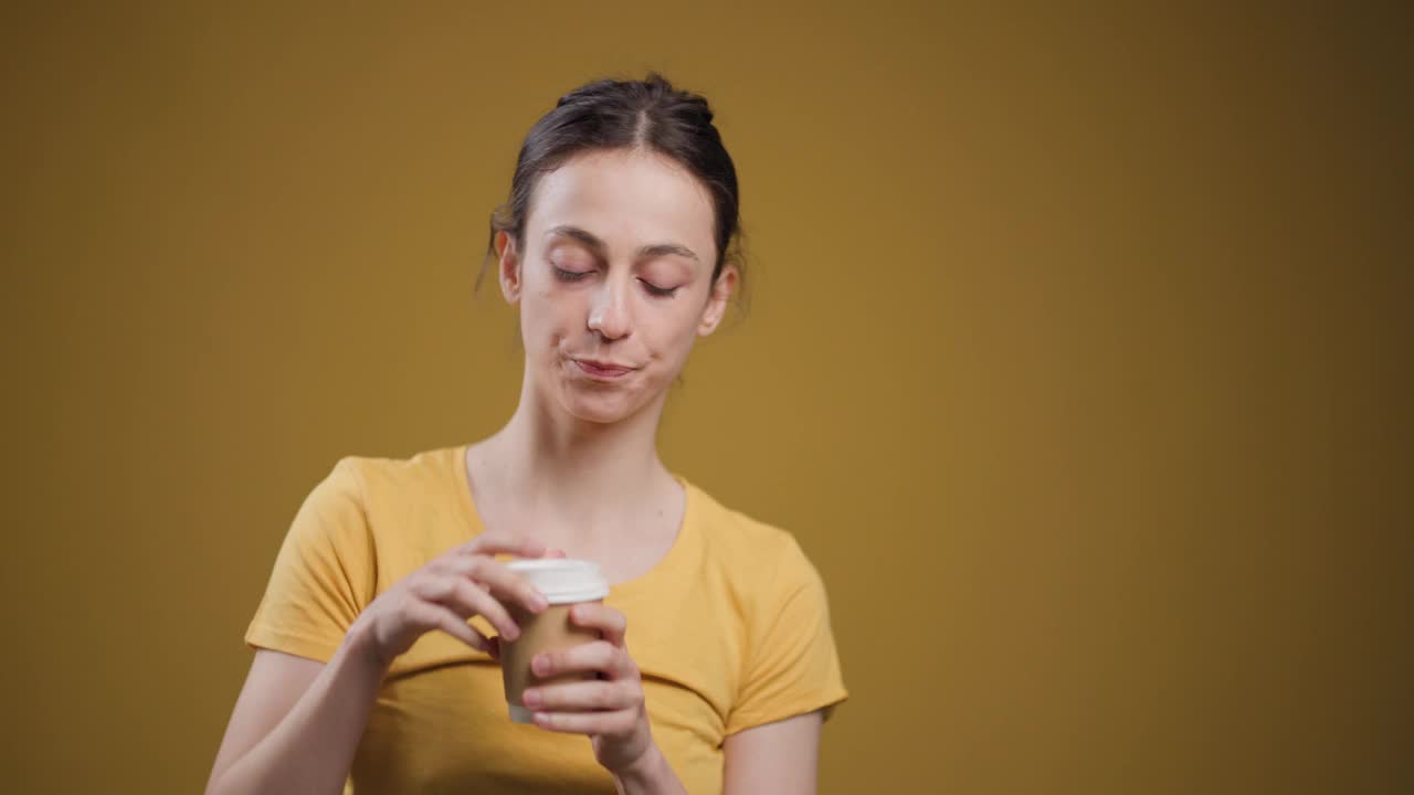 一个沉思的年轻女子从一个一次性杯子里品尝咖啡的香气，背景是芥末黄色。视频素材