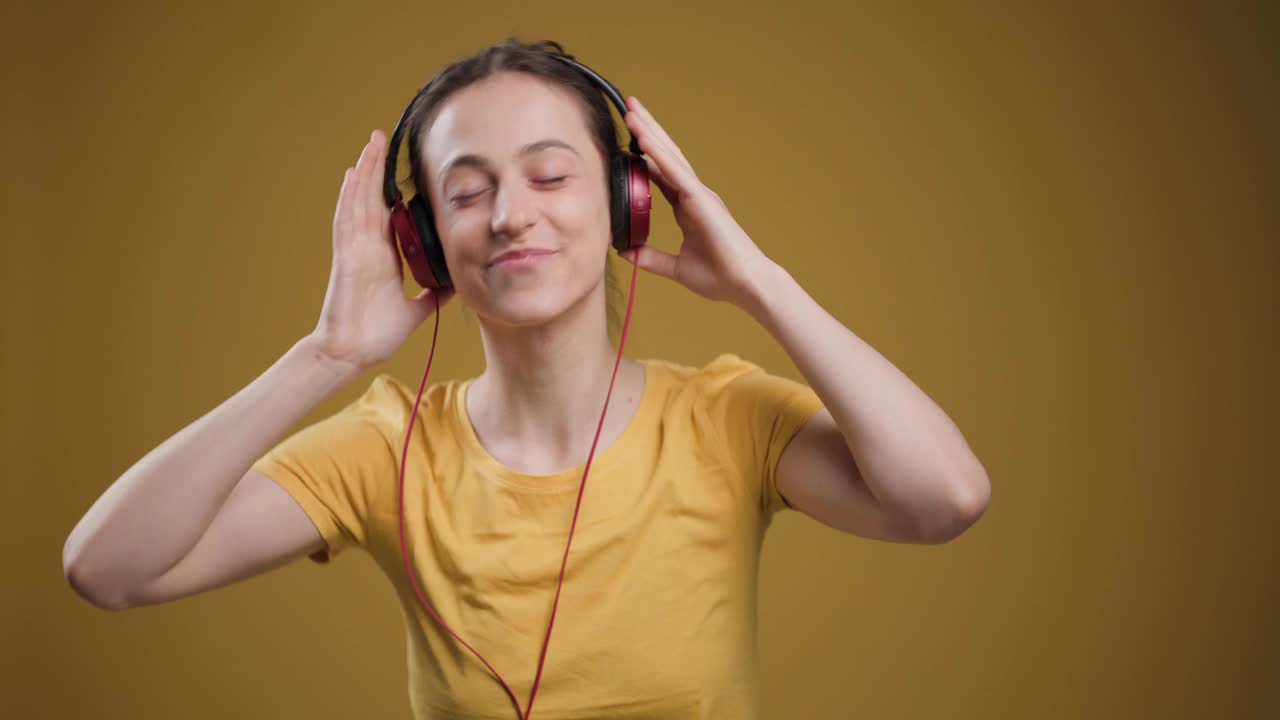 一个快乐的年轻女子闭上眼睛，戴着耳机听音乐，在黄色的背景下散发着快乐和放松。视频素材